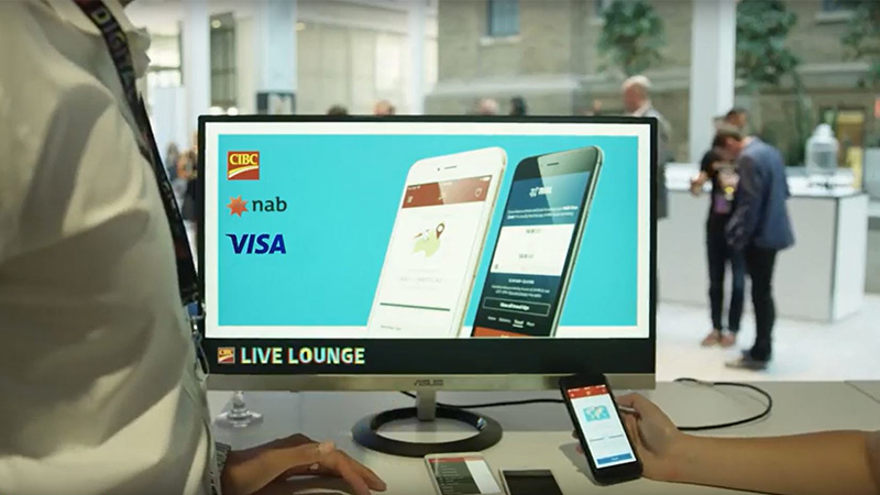 Two people talk in front of a computer screen showing Visa, CIBC, Nab logos and two phones in the CIBC Live Lounge. 